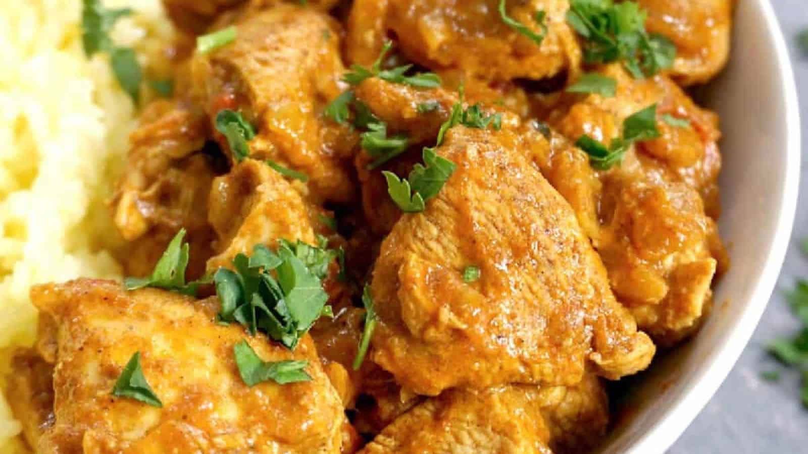 A bowl containing chunks of chicken in a thick orange sauce, garnished with chopped cilantro, next to a serving of yellow rice.