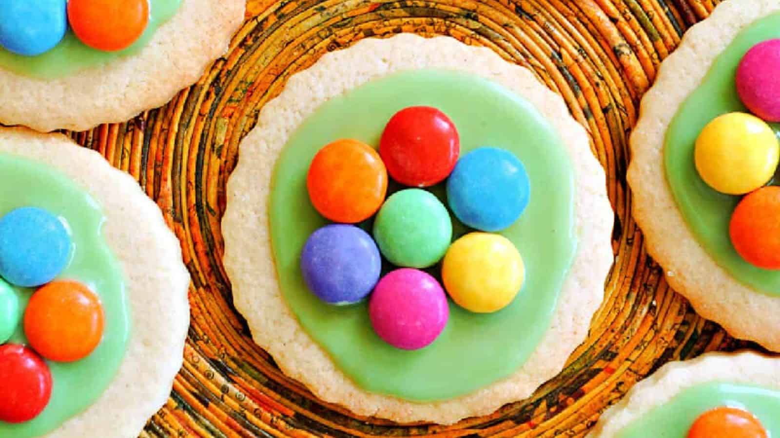 Round cookies with green icing, each topped with a cluster of colorful chocolate candies, arranged on a woven multicolored surface.