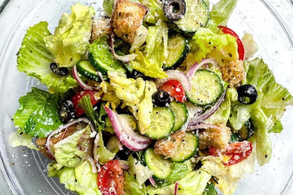 A bowl containing lettuce, cucumber slices, tomato wedges, red onion rings, black olives, croutons, and grated cheese.