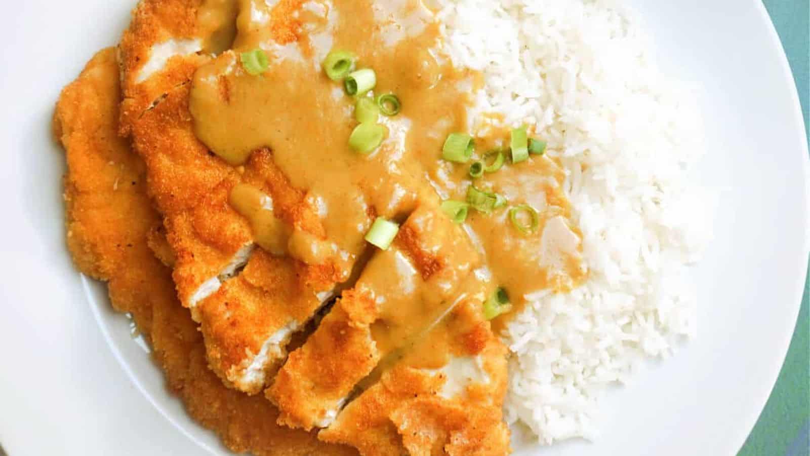 A plate with sliced breaded chicken cutlet topped with brown sauce and chopped green onions, served next to white rice.
