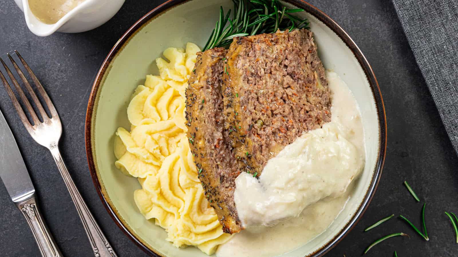 Slices of meatloaf, mashed potatoes topped with white gravy and a sprig of rosemary arranged on a plate; beside the plate are a fork, knife, and a small sauce pitcher.
