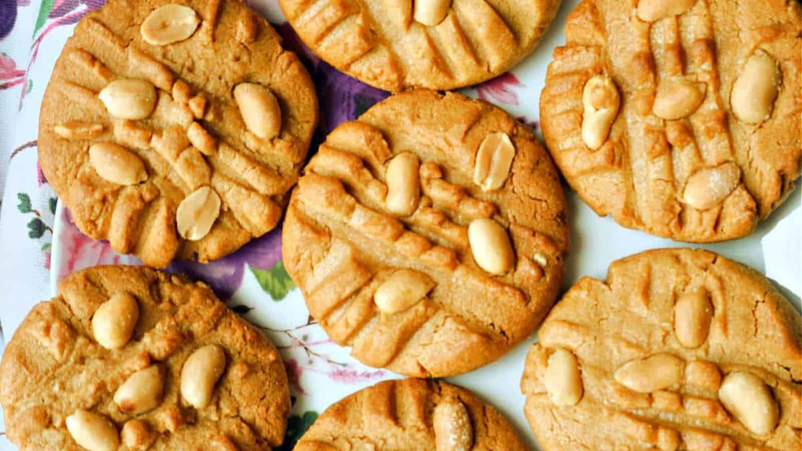 Seven peanut butter cookies with crisscross fork marks and whole peanuts on top, placed on a plate with a floral pattern.