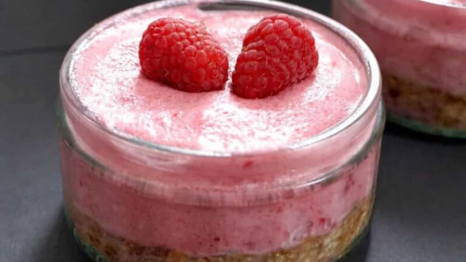 A glass jar containing a layered dessert with a crumbly base, pink mousse topping, and two raspberries placed on top.