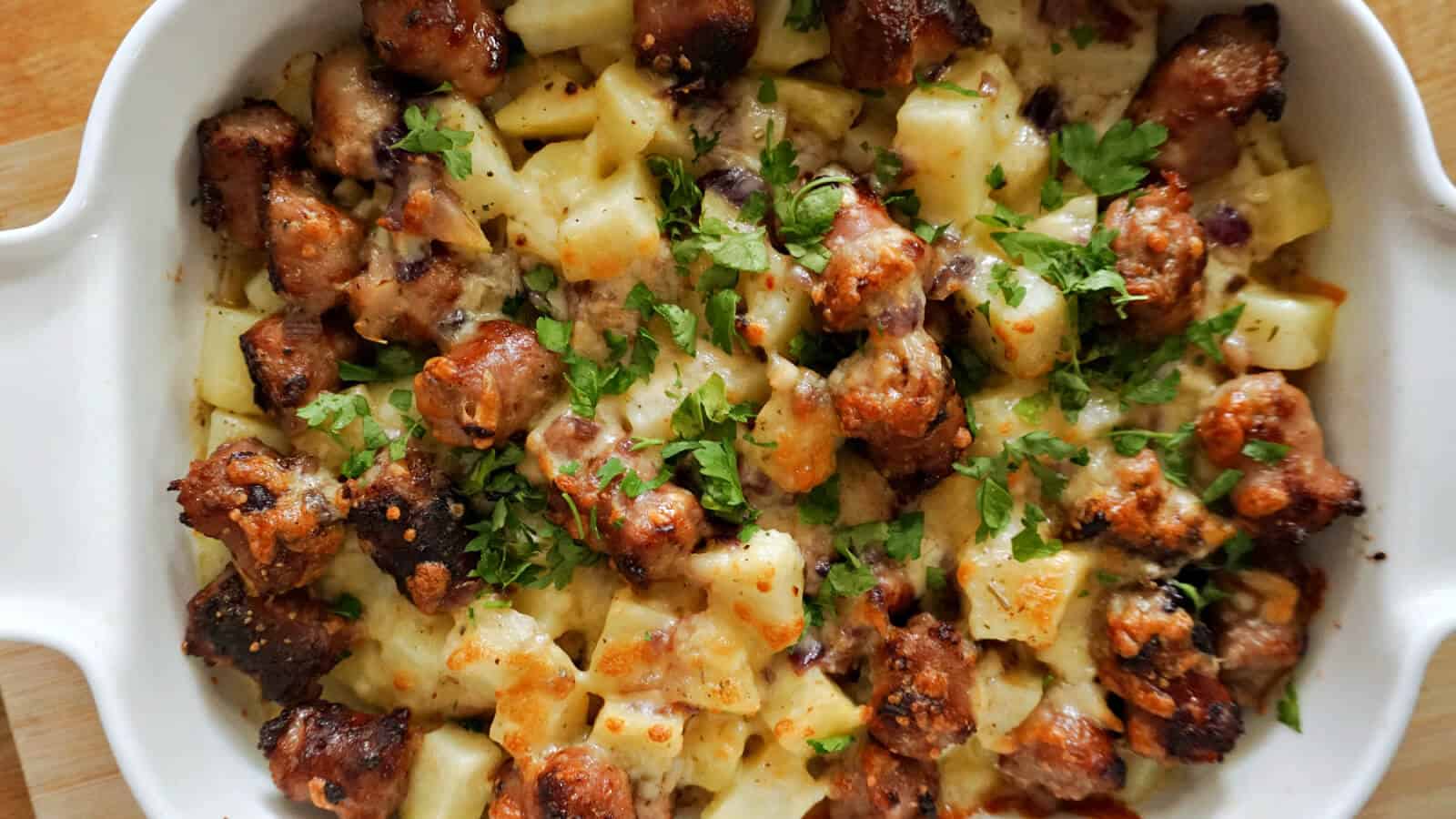 A baked casserole dish with browned sausage pieces, diced potatoes, melted cheese, and chopped parsley on top.