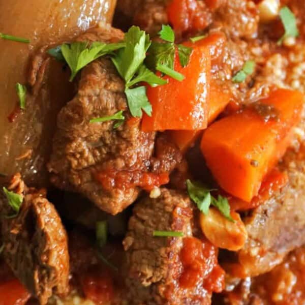 Chunks of beef, carrots, onions, and tomatoes in a stew, garnished with fresh parsley and served over couscous.