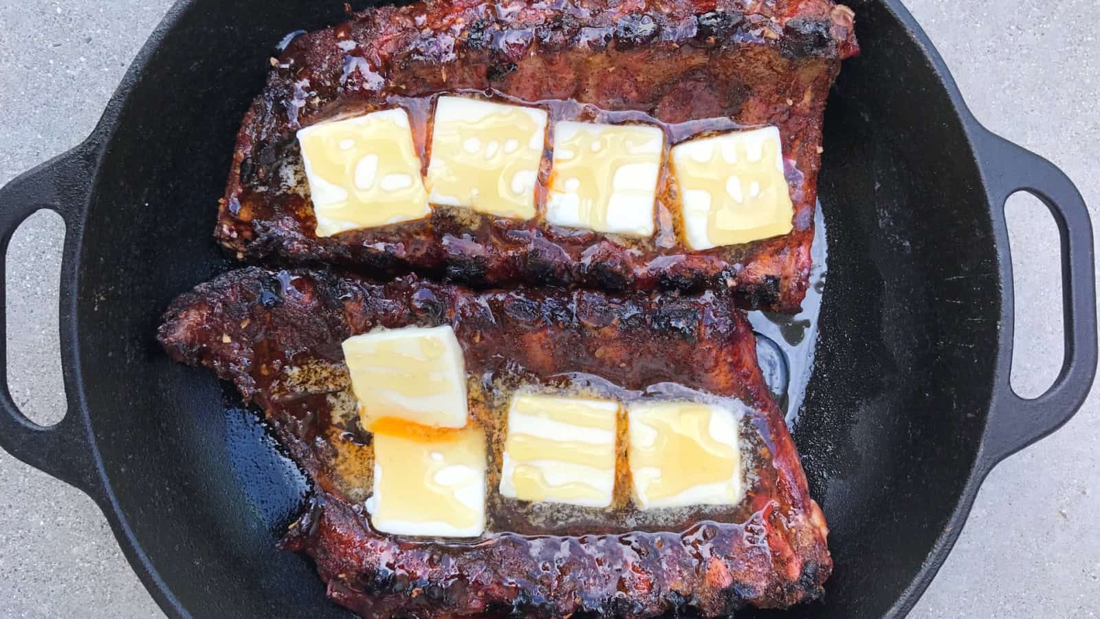 Two racks of grilled ribs in a cast iron pan, each topped with slices of butter and drizzled with honey.