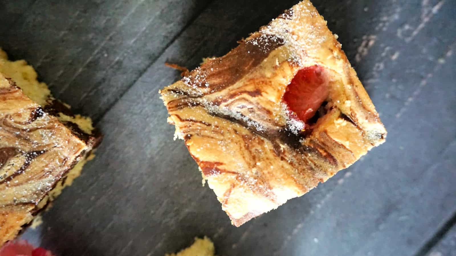 A square piece of marbled cake with a visible raspberry on top, placed on a dark surface.