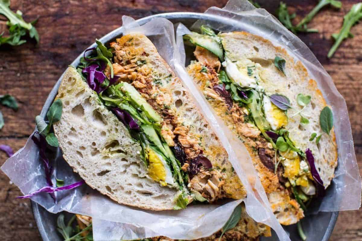 A close-up of a plate holding two halves of a rustic sandwich, filled with greens and shredded meat, wrapped in parchment paper and placed on a wooden surface.