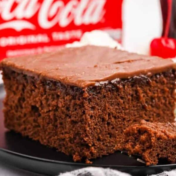 A slice of chocolate cake with chocolate frosting sits on a black plate. In the background, there is a cherry and a dollop of whipped cream.