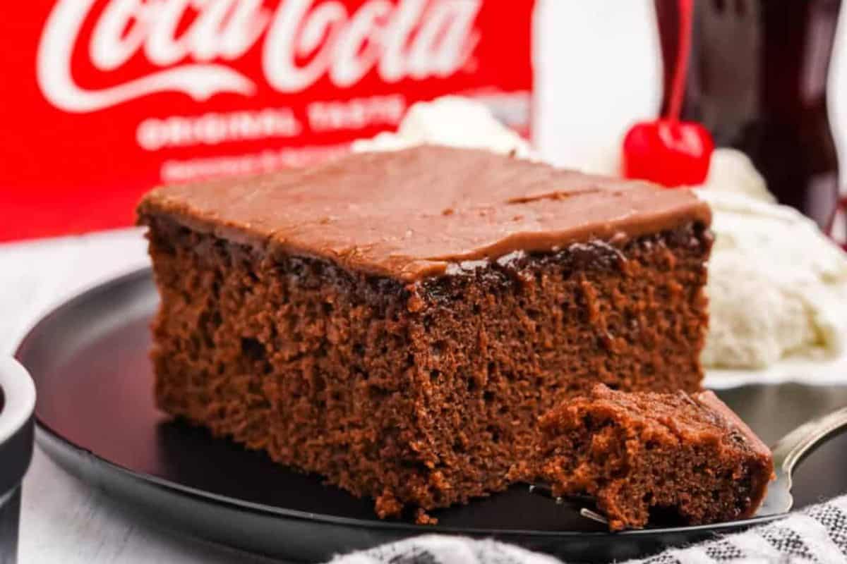 A slice of chocolate cake with thick frosting rests on a black plate, accompanied by a scoop of vanilla ice cream and a cherry. In the background, there is a classic Coca-Cola sign.
