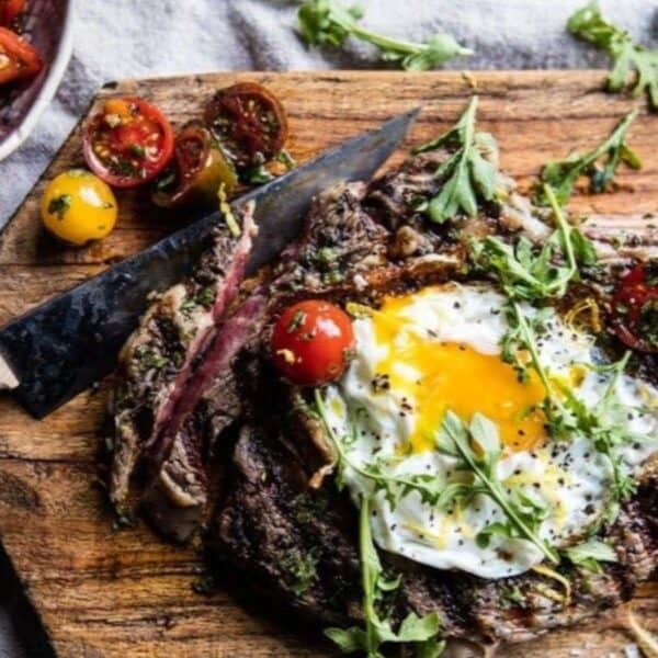 A grilled steak topped with a sunny-side-up egg, cherry tomatoes, and herbs on a wooden board, accompanied by a small bowl of salt and a colorful tomato salad in a separate bowl.