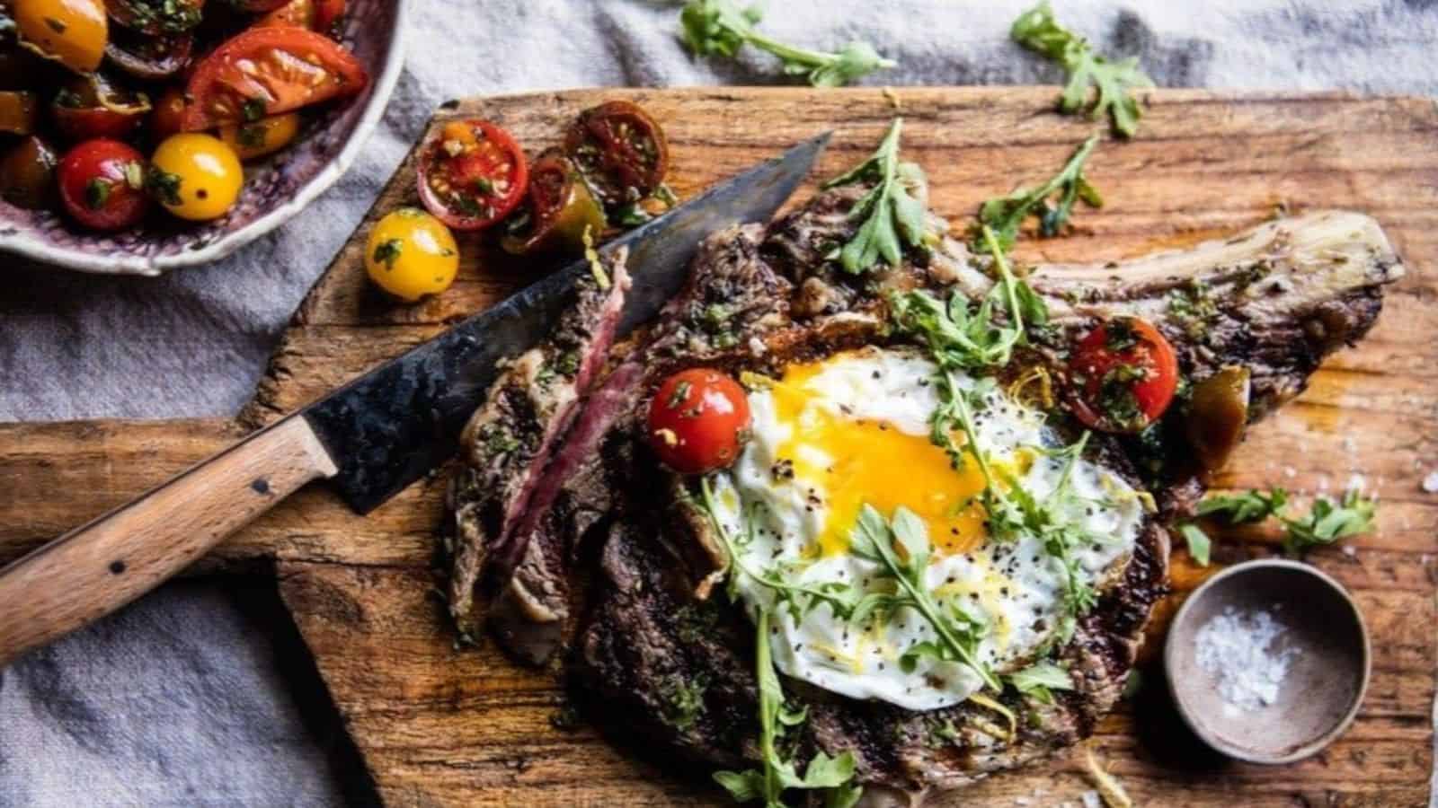 A grilled steak topped with a sunny-side-up egg, cherry tomatoes, and herbs on a wooden board, accompanied by a small bowl of salt and a colorful tomato salad in a separate bowl.
