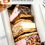 Three Neapolitan waffle ice cream sandwiches, each coated with a different topping, are arranged in a parchment-lined container on a marble surface.