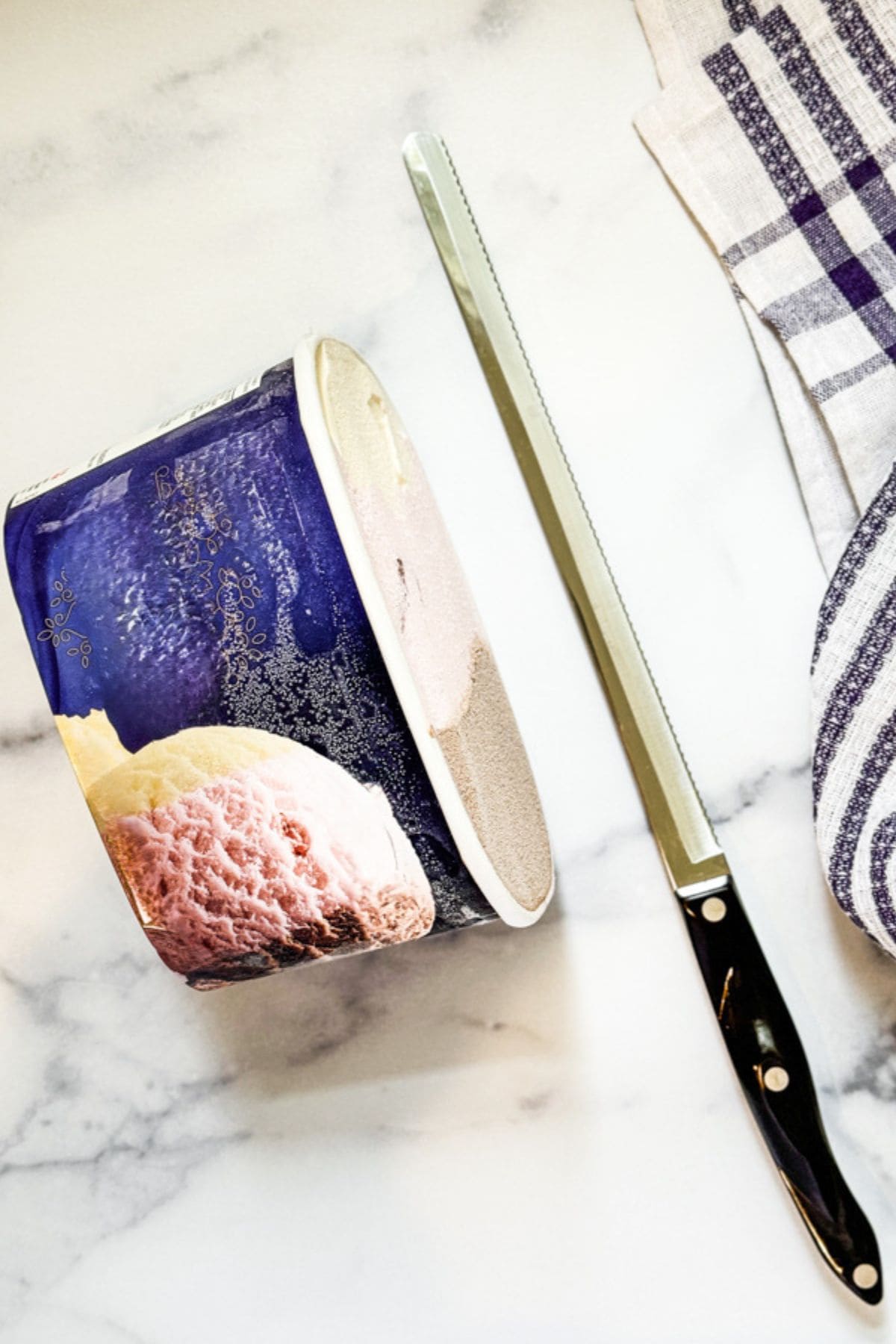 A half-cut ice cream container, a long serrated knife, and a striped kitchen towel rest on a marble surface.
