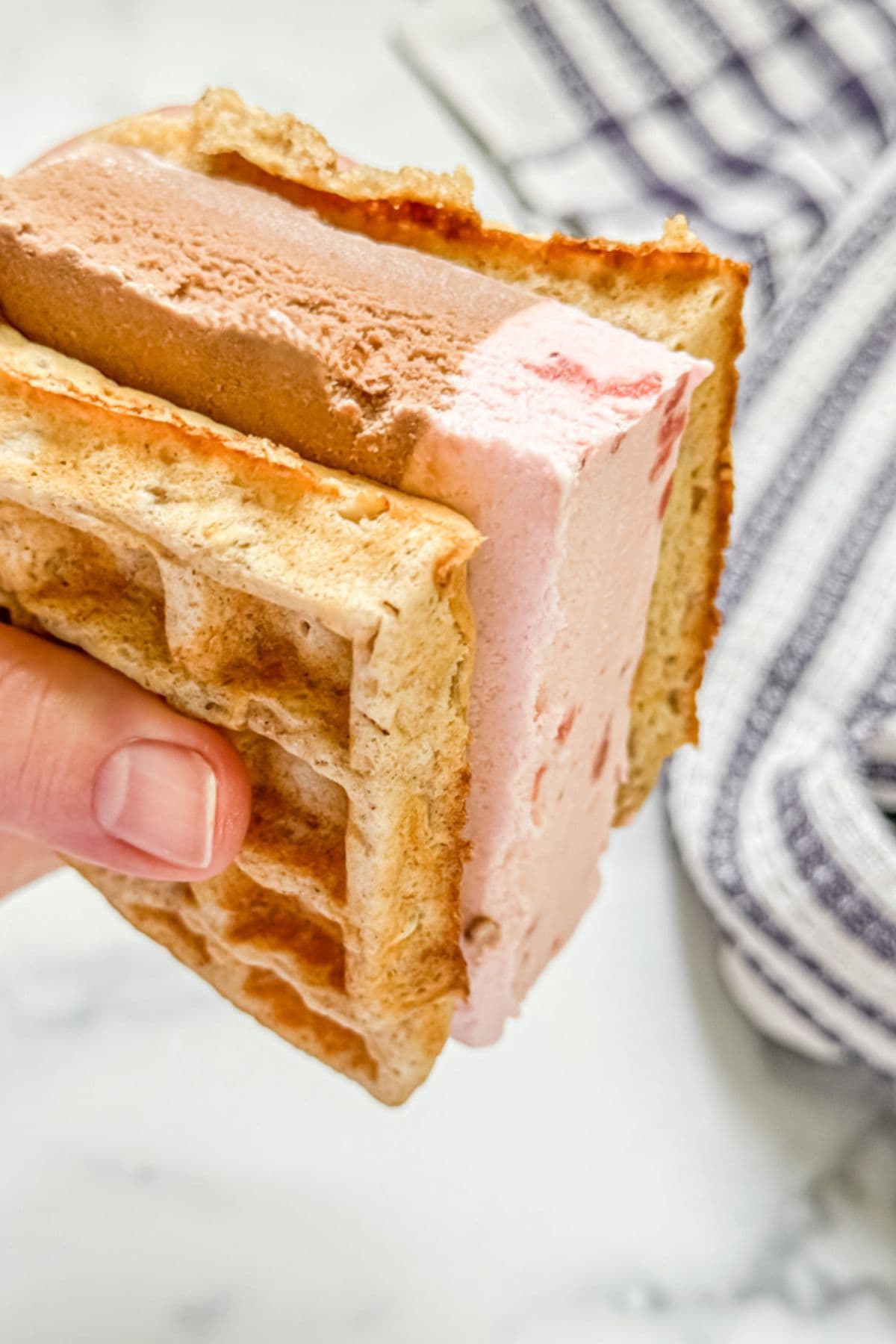 A hand holds a waffle ice cream sandwich with layers of chocolate and strawberry ice cream; a striped cloth is in the background.