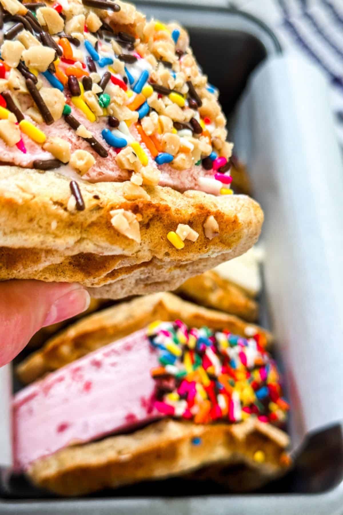 A close-up of waffle ice cream sandwiches filled with colorful sprinkles and nuts, held above a tray with more sandwiches.