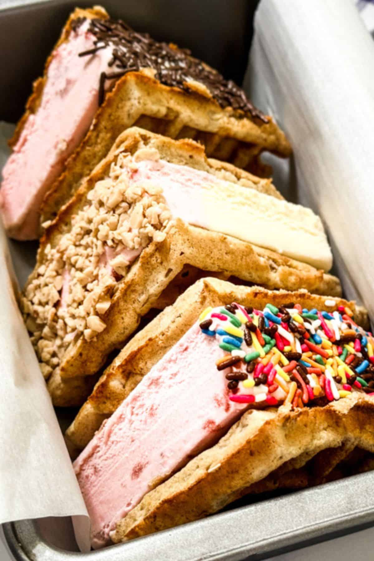 Three waffle ice cream sandwiches with different ice cream flavors, each topped with chocolate sprinkles, chopped nuts, or rainbow sprinkles, arranged in a metal tray lined with parchment paper.