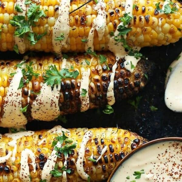 Three grilled corn cobs topped with creamy sauce and chopped herbs, accompanied by a bowl of extra sauce and a spoon.