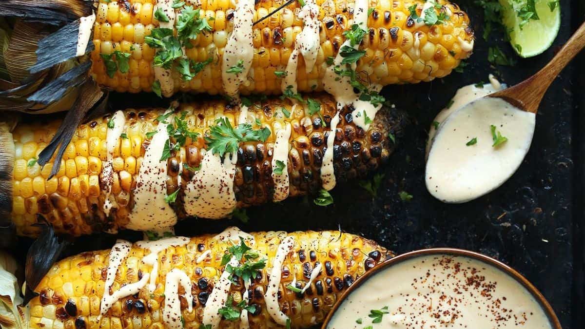 Three grilled corn cobs topped with creamy sauce and chopped herbs, accompanied by a bowl of extra sauce and a spoon.