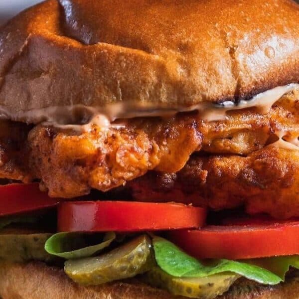 A toasted bun filled with fried chicken, lettuce, tomato slices, pickles, and sauce.