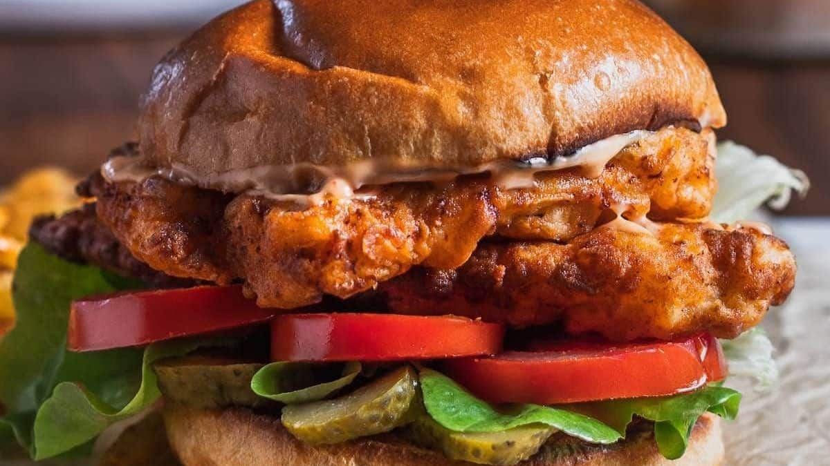 A toasted bun filled with fried chicken, lettuce, tomato slices, pickles, and sauce.