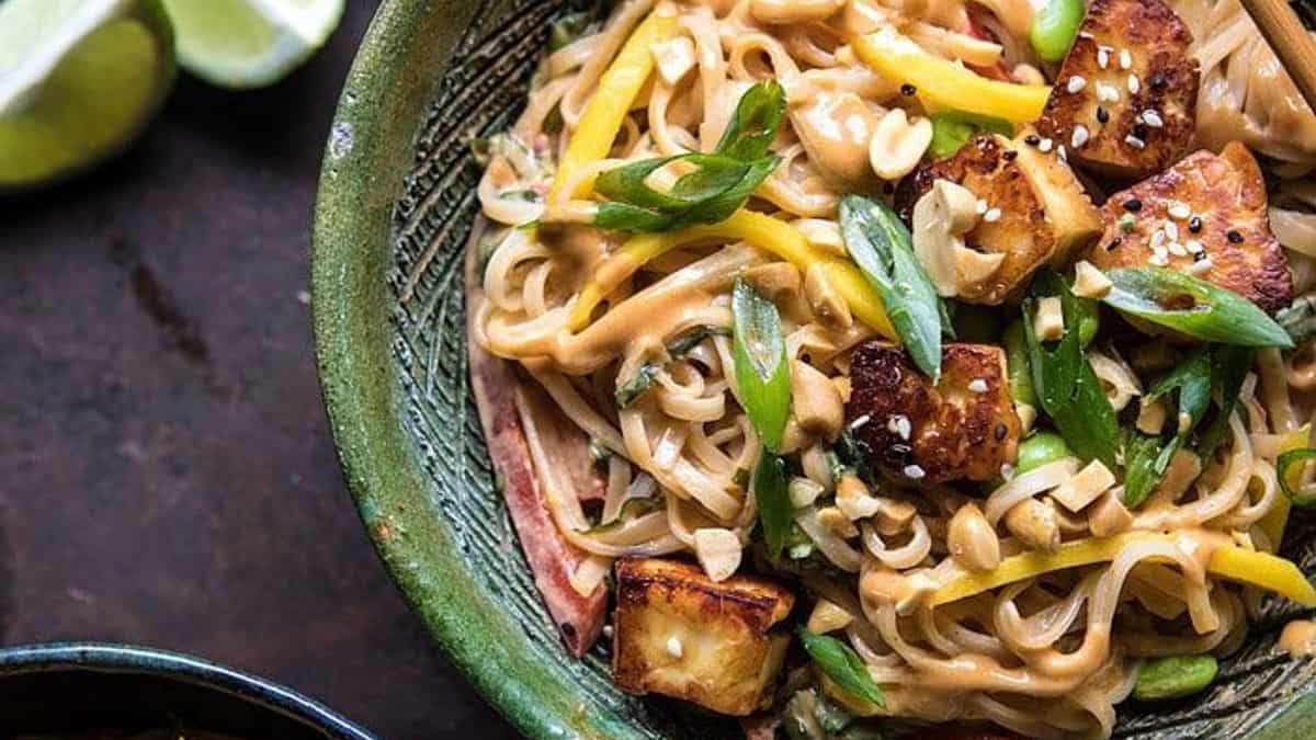 A bowl of noodle salad with tofu cubes, sliced green onions, yellow peppers, peanuts, and a creamy sauce; lime wedges are in the background.