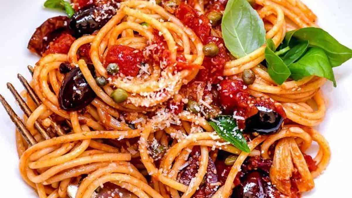 A plate of spaghetti topped with tomato sauce, black olives, capers, grated cheese, garlic slices, and fresh basil, with a fork placed beside it.