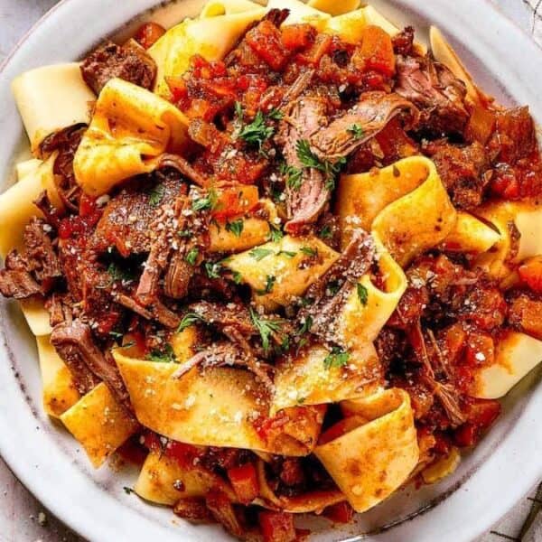 Wide ribbons of pappardelle pasta topped with shredded beef ragu and garnished with chopped herbs on a plate, accompanied by a cloth napkin and sprigs of fresh parsley.