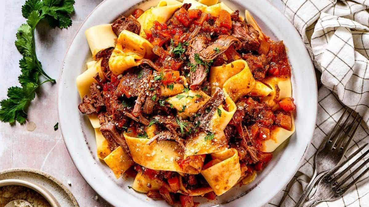 Wide ribbons of pappardelle pasta topped with shredded beef ragu and garnished with chopped herbs on a plate, accompanied by a cloth napkin and sprigs of fresh parsley.