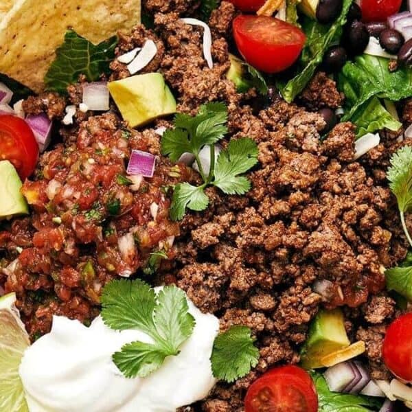 Ground beef, black beans, avocado slices, cherry tomatoes, lettuce, shredded cheese, tortilla chips, salsa, sour cream, red onion pieces, cilantro leaves, and a lime wedge.
