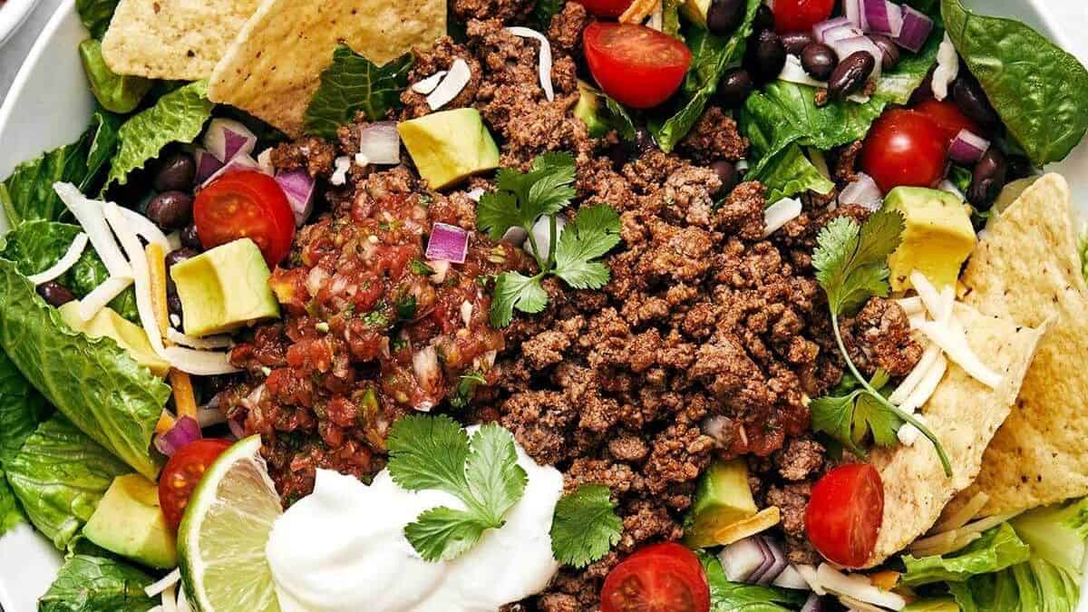 Ground beef, black beans, avocado slices, cherry tomatoes, lettuce, shredded cheese, tortilla chips, salsa, sour cream, red onion pieces, cilantro leaves, and a lime wedge.