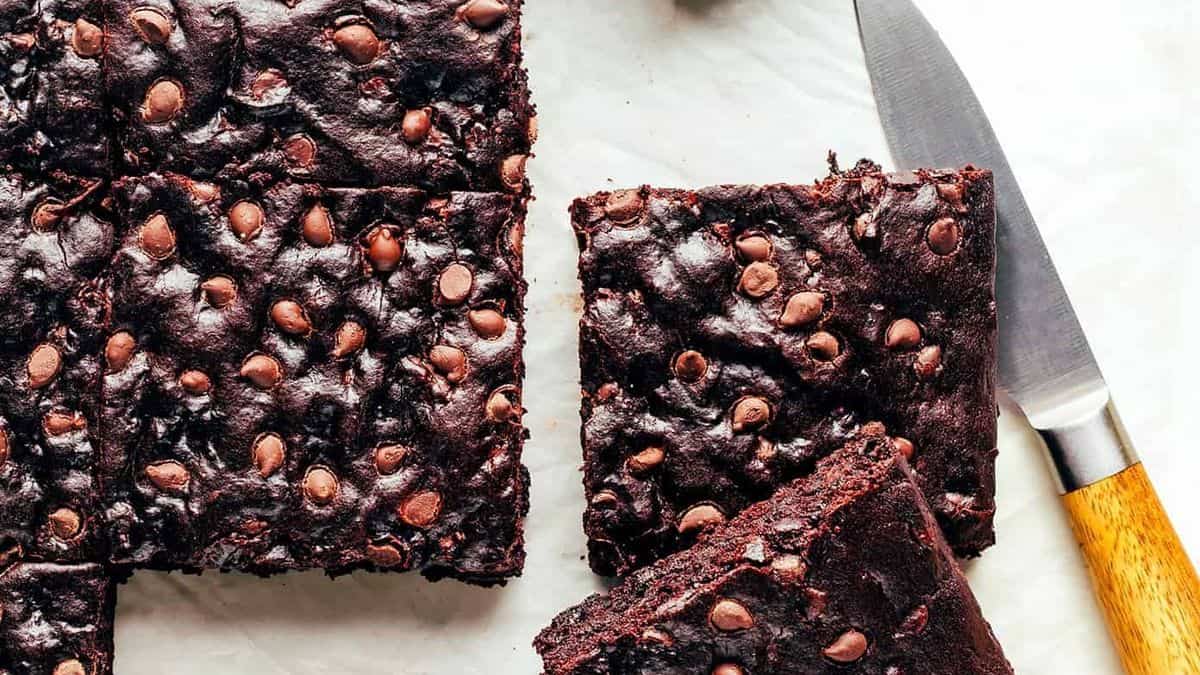 Squares of chocolate chip brownies arranged on a white surface, with a knife positioned beside them.