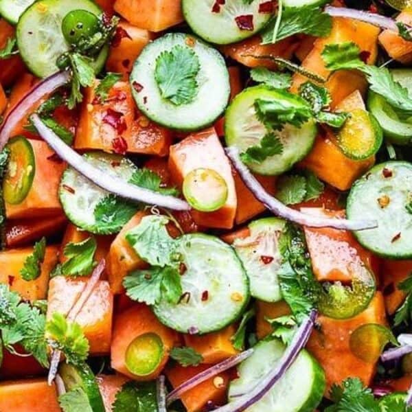 Cubed papaya, sliced cucumber, red onion, cilantro, baby greens, and red pepper flakes in a white bowl.