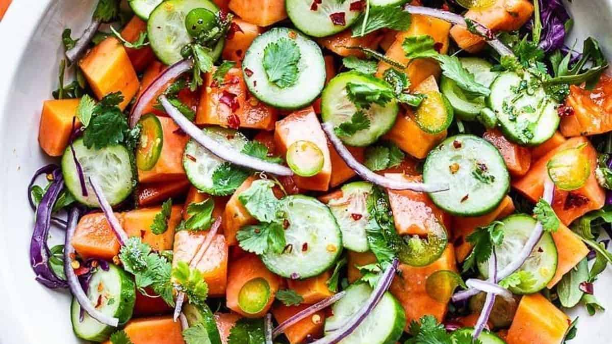 Cubed papaya, sliced cucumber, red onion, cilantro, baby greens, and red pepper flakes in a white bowl.