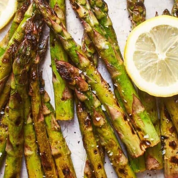 Several grilled asparagus spears, topped with coarse salt and cracked pepper, are arranged with fresh lemon slices on a white background.