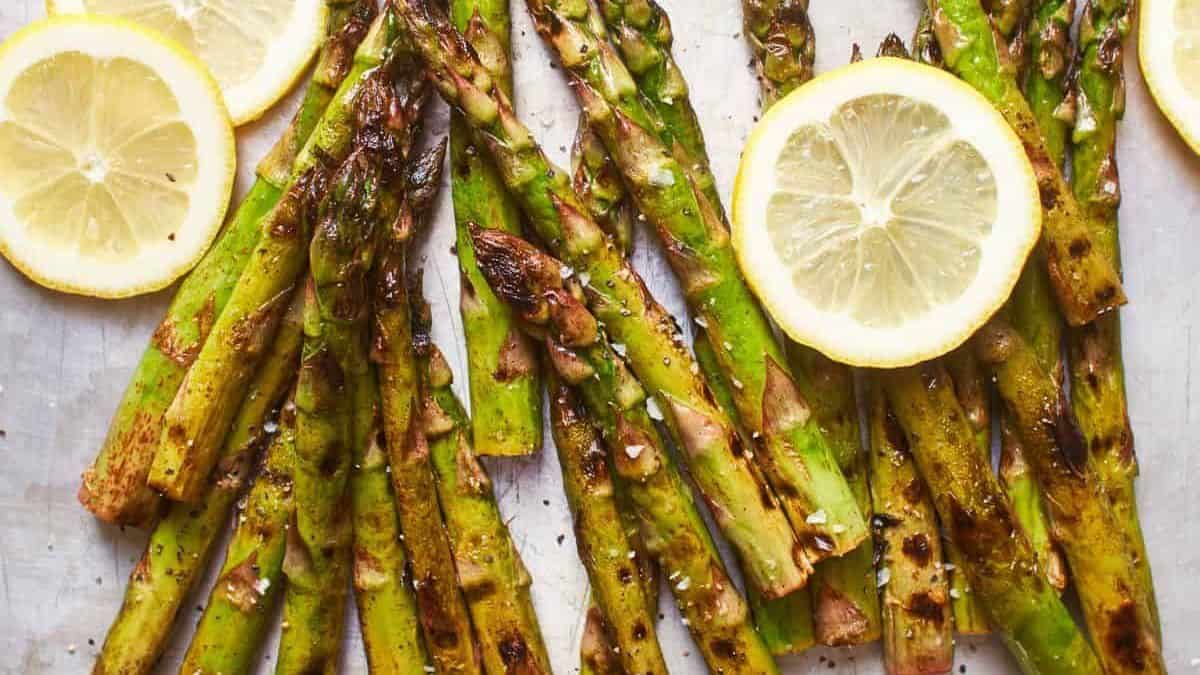 Several grilled asparagus spears, topped with coarse salt and cracked pepper, are arranged with fresh lemon slices on a white background.