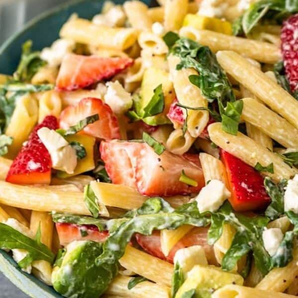 A bowl containing penne pasta, sliced strawberries, avocado pieces, spinach leaves, feta cheese crumbles, and fresh basil.