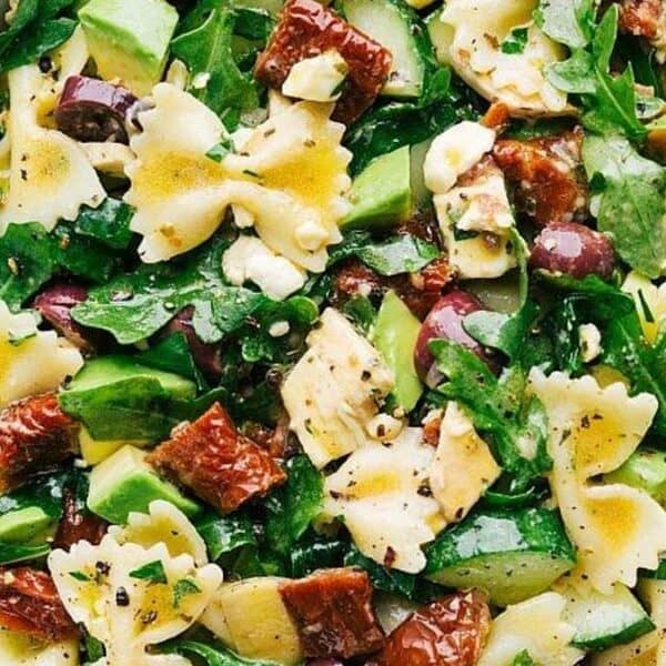 A bowl containing bowtie pasta, sun-dried tomatoes, avocado pieces, feta cheese cubes, black olives, arugula leaves, and vinaigrette dressing; a spoon is placed next to the bowl.