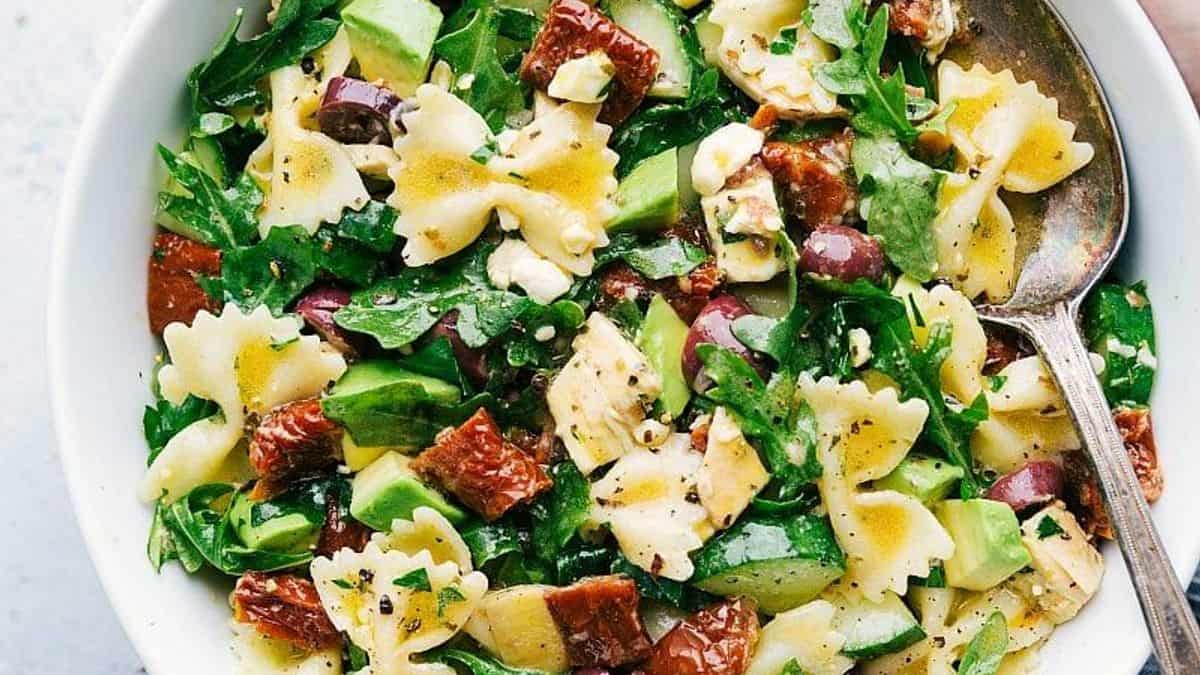 A bowl containing bowtie pasta, sun-dried tomatoes, avocado pieces, feta cheese cubes, black olives, arugula leaves, and vinaigrette dressing; a spoon is placed next to the bowl.