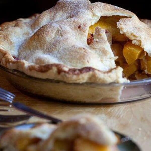 A baked peach pie with a golden, flaky crust sits on a wooden surface. One slice has been removed, showing the juicy fruit filling inside.