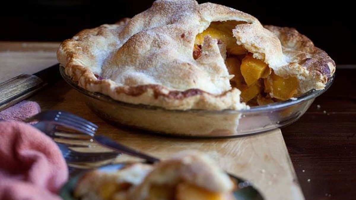 A baked peach pie with a golden, flaky crust sits on a wooden surface. One slice has been removed, showing the juicy fruit filling inside.