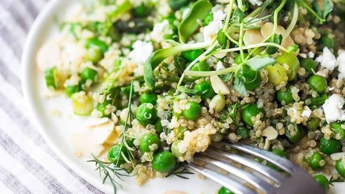 A plate containing quinoa, green peas, sliced green onions, leafy greens, herbs, and crumbled cheese; a fork rests on the plate.