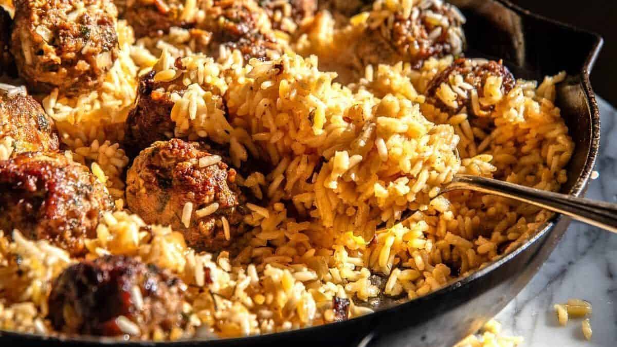A skillet filled with yellow rice and browned meatballs, with a spoon scooping up the rice.