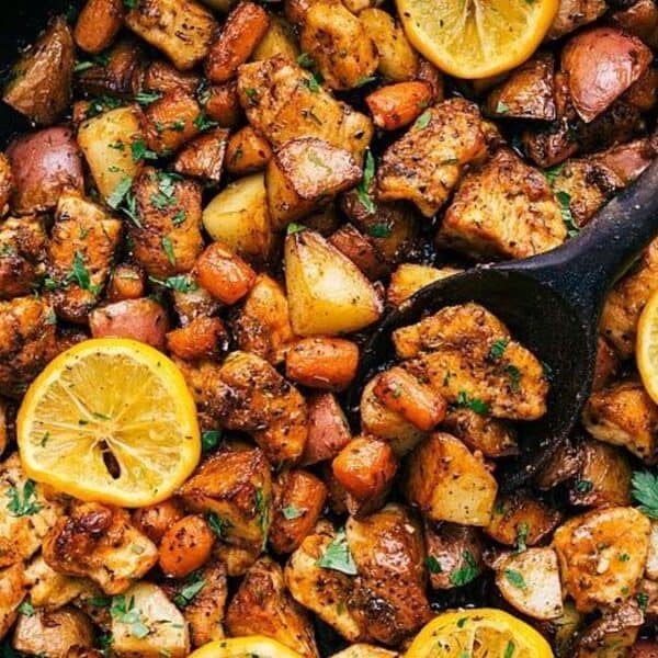 A skillet containing chicken pieces, diced potatoes, carrots, lemon slices, and fresh herbs, with a wooden spoon inside.