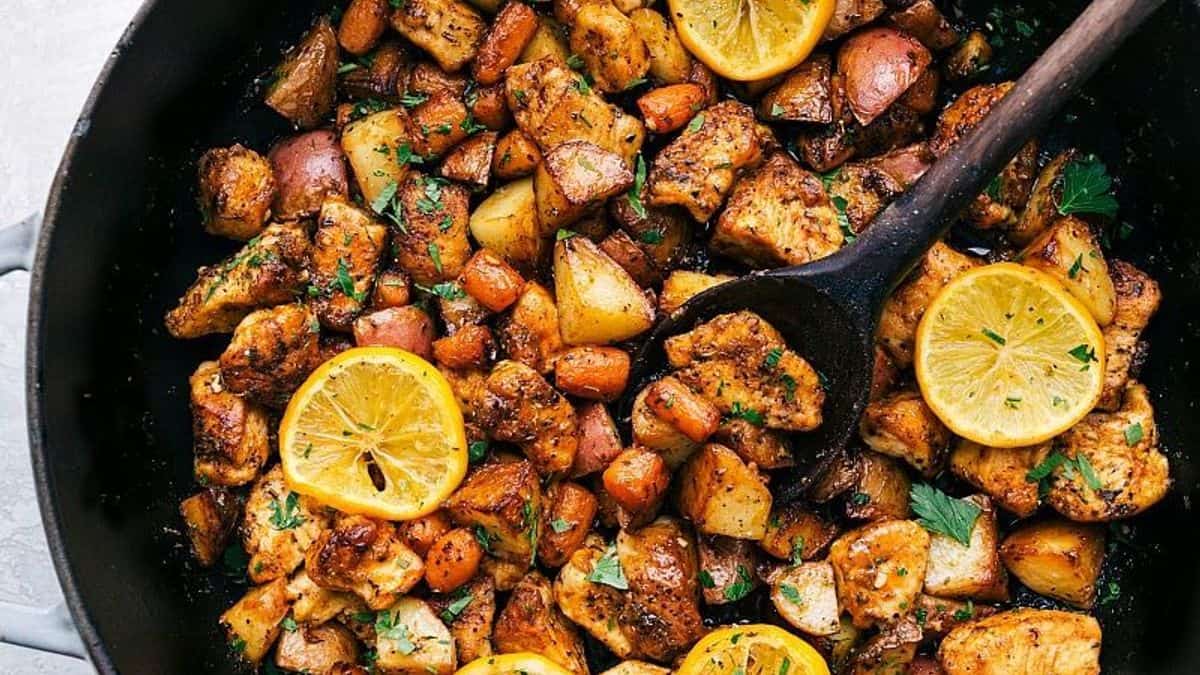 A skillet containing chicken pieces, diced potatoes, carrots, lemon slices, and fresh herbs, with a wooden spoon inside.