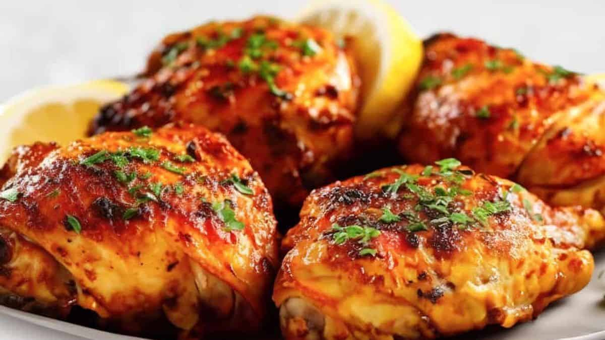 Four pieces of roasted chicken garnished with chopped herbs are arranged on a plate, with lemon wedges in the background.
