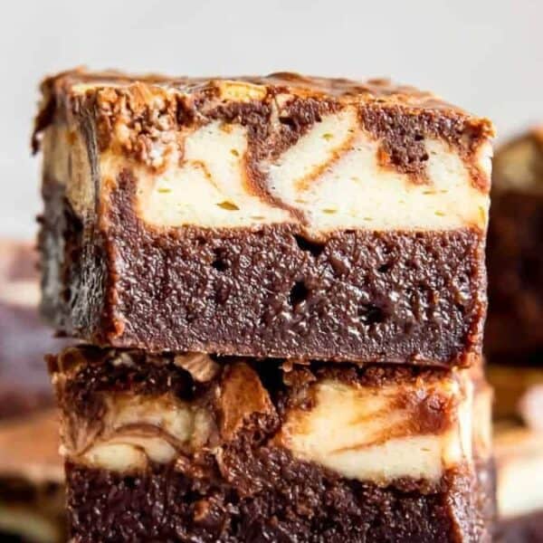 Two cheesecake swirl brownie bars stacked, showing a dark, fudgy base and marbled cheesecake layer, with additional brownies scattered in the background.