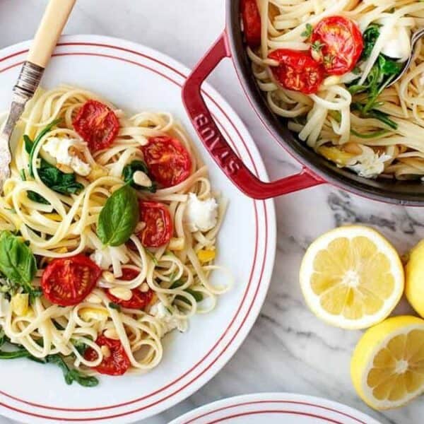 A plate and pot of linguine pasta with tomatoes, spinach, basil, and cheese on a marble surface alongside fresh tomatoes and sliced lemons.