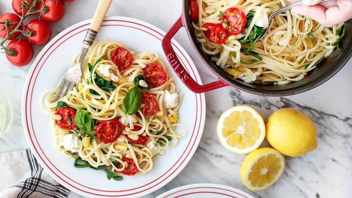 A plate and pot of linguine pasta with tomatoes, spinach, basil, and cheese on a marble surface alongside fresh tomatoes and sliced lemons.