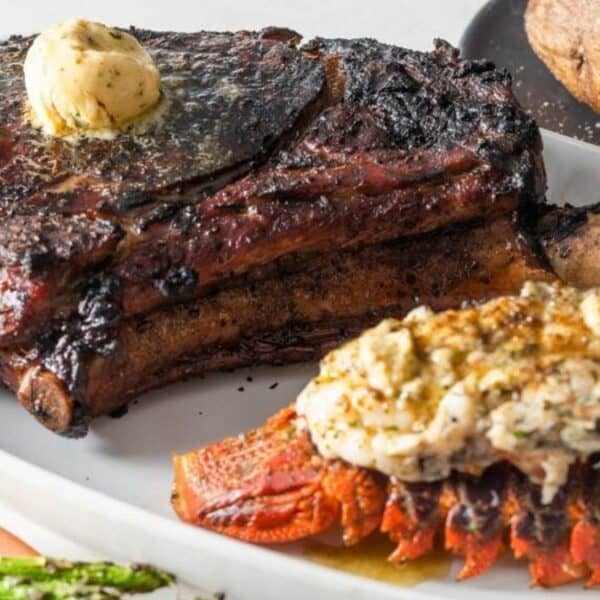A grilled tomahawk steak topped with herb butter, served alongside a broiled lobster tail, baked potato, and asparagus.